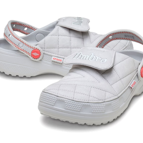 Beyaz, kroko desenli Crocs ayakkabı. Turuncu düğme detayları ve markalı bantlar mevcut. "Umbro" ve "Crocs" logoları görünüyor.