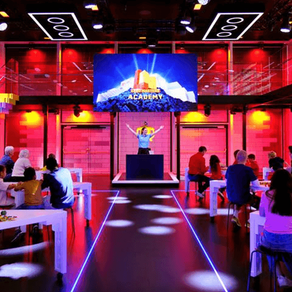 Kırmızı aydınlatmalı bir odada çocuklar ve yetişkinler masalarda LEGO yapıyor. Arkada "ACADEMY" yazılı ekran. Eğlenceli atmosfer.