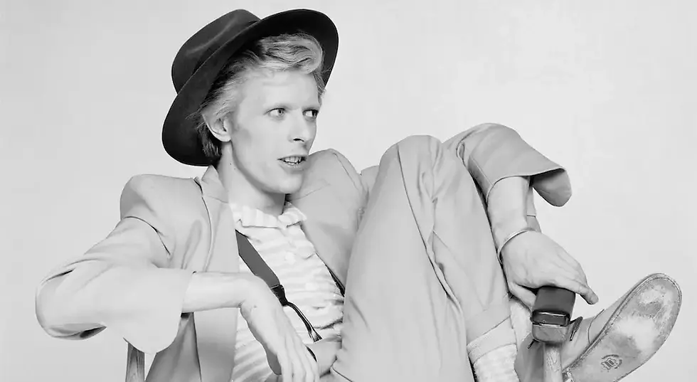 Bir adam şapka takıyor, soluk renkli takım elbise giymiş, boş bir fon önünde rahatça oturuyor, düşünceli bir ifadeye sahip.