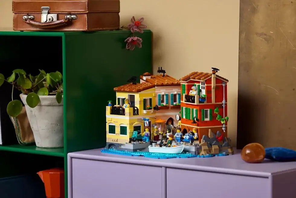 Lego kasaba maketi, deniz kenarında, renkli binalar ve minifigürlerle yerleştirilmiş. Yeşil raf, kahverengi bavul ve bitki eşlik ediyor.