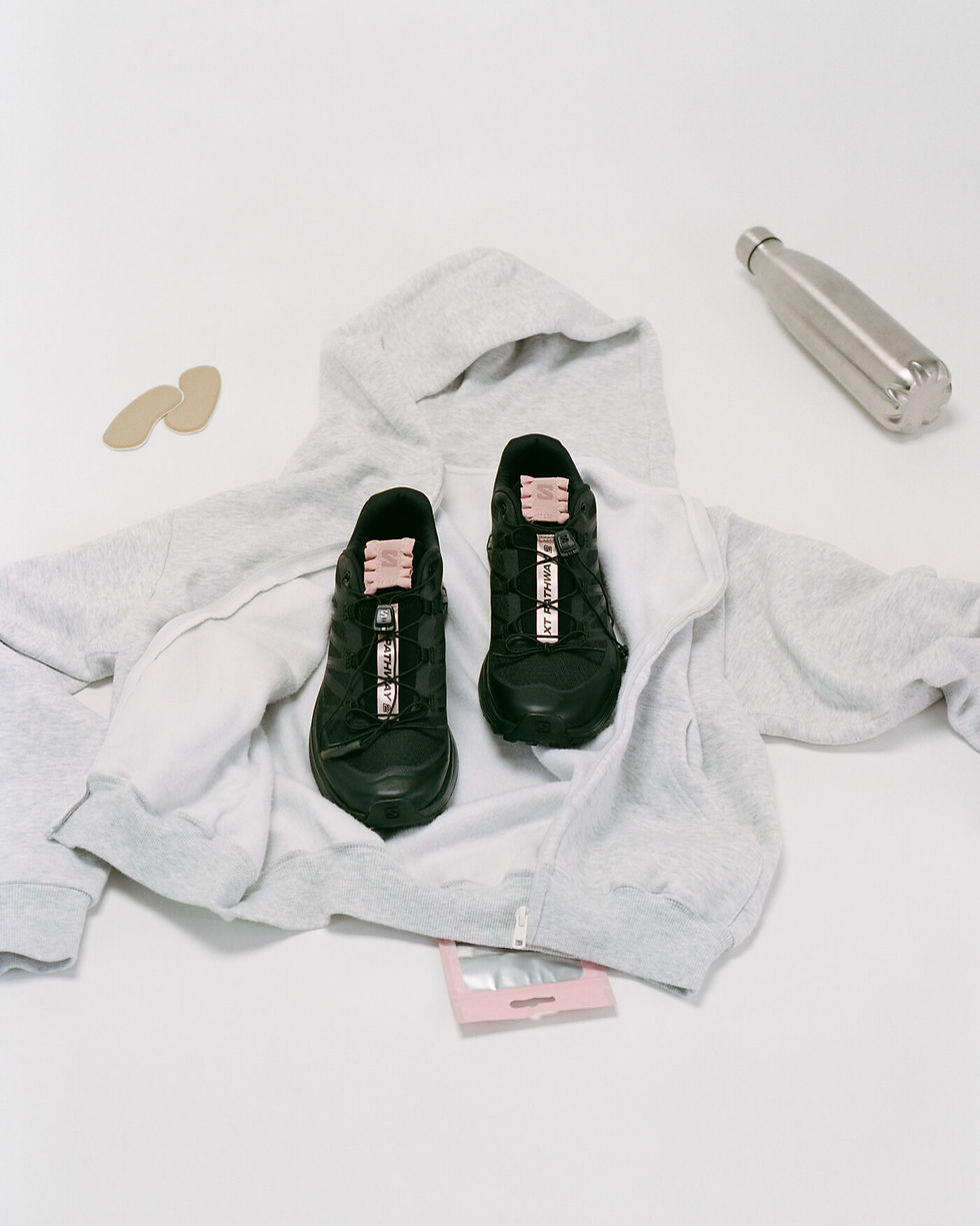 Gri zeminde gri kapüşonlu üst, siyah spor ayakkabılar, metal su şişesi ve ten rengi pedler yer alıyor. Ayakkabılarda pembe diller var.