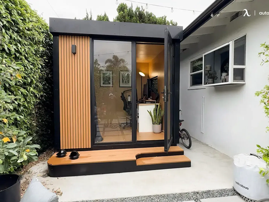 Small wooden shed with glass door, black trim, and open interior showing a desk. Surrounded by greenery, bike nearby, calm setting.