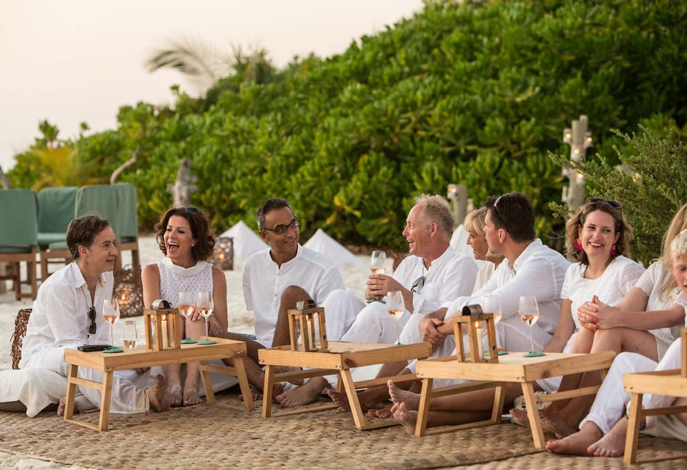 Grupo de umas 9 pessoas sentadas em um tapete bege sobre a areia. Todos estão descalços, vestidos de branco, com expressões felizes, tomando champanhe em um fim de tarde nas Maldivas