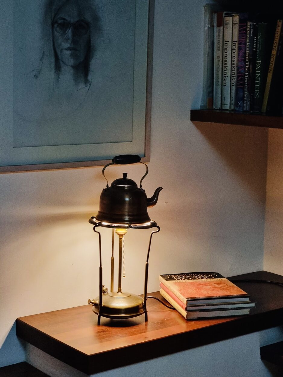 An old oil cooker with a kettle on top, made into a table lamp, lit on a wooden shelf