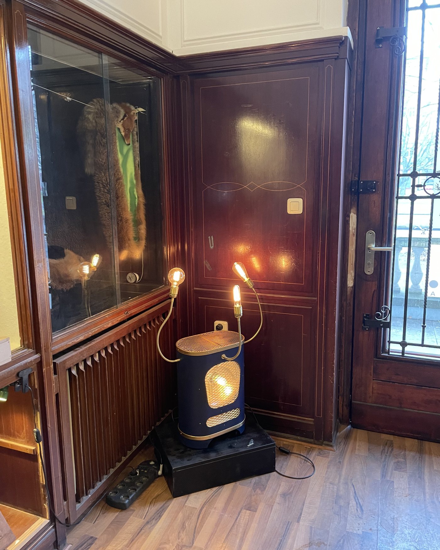 Blue Heater lamp lit on the floor of a wood-panelled entrance hall, beside a glass door