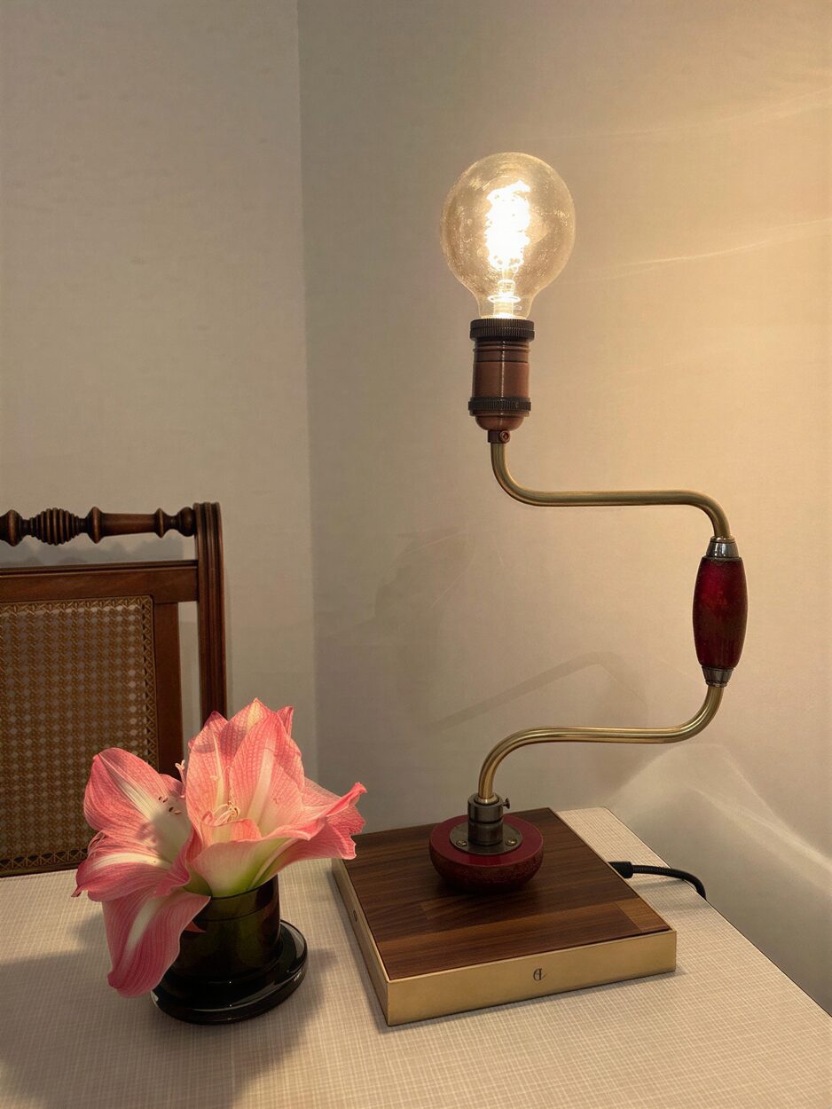 A hand brace drill made into a table lamp with one bulb, on a walnut and brass base beside pink flowers