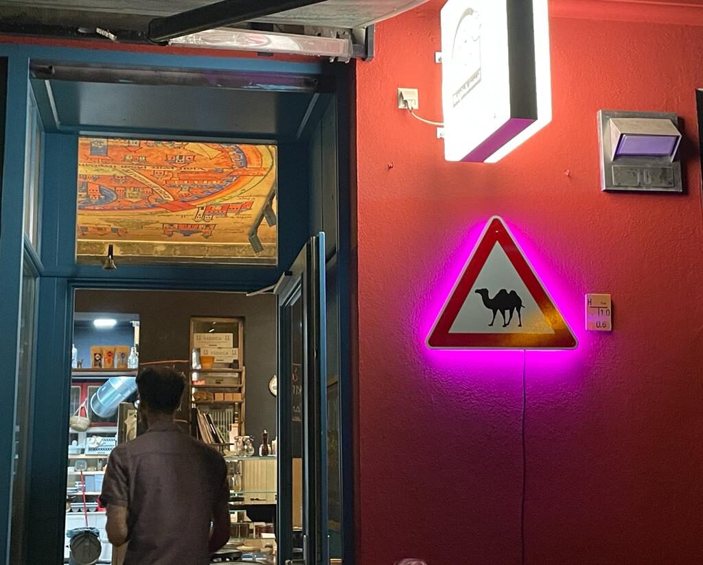 A triangular camel warning sign glowing pink on the red wall beside the open restaurant door at night