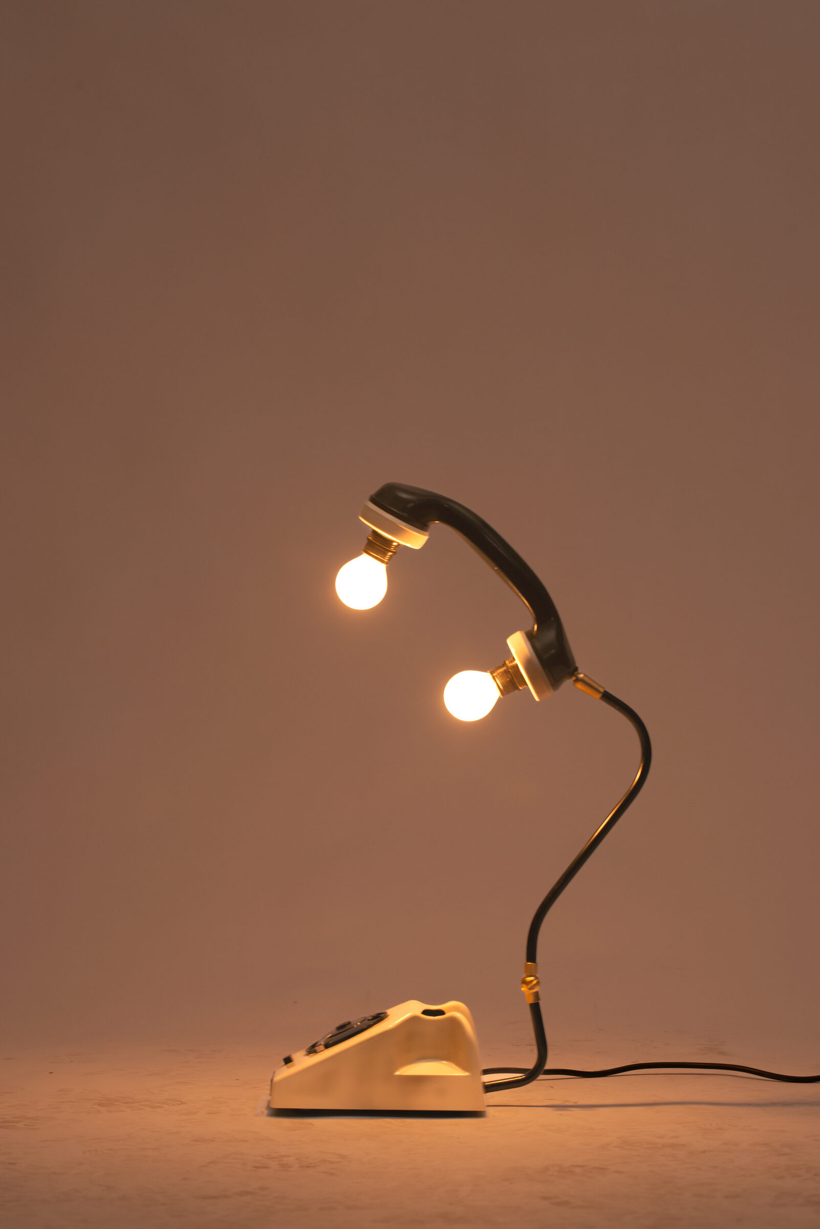 A cream rotary telephone made into a table lamp, two bulbs lit on a curved brass arm