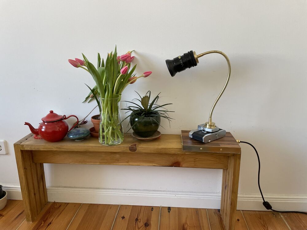 An analogue camera on a bent brass arm, made into a table lamp, on a wooden bench with flowers