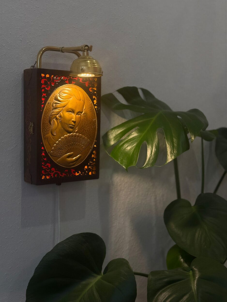 A wooden jewellery box with a brass portrait relief, made into a lit wall lamp beside a monstera plant