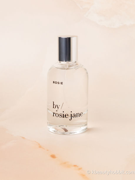 Perfume bottle labeled "ROSIE by/rosie jane" on a light beige marbled background. The mood is elegant and minimalistic.