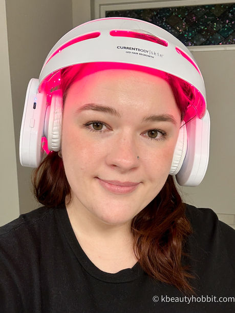 Woman wearing a white LED hair regrowth helmet emitting red light, labeled "CURRENTBODY Skin." Neutral expression, indoor setting.