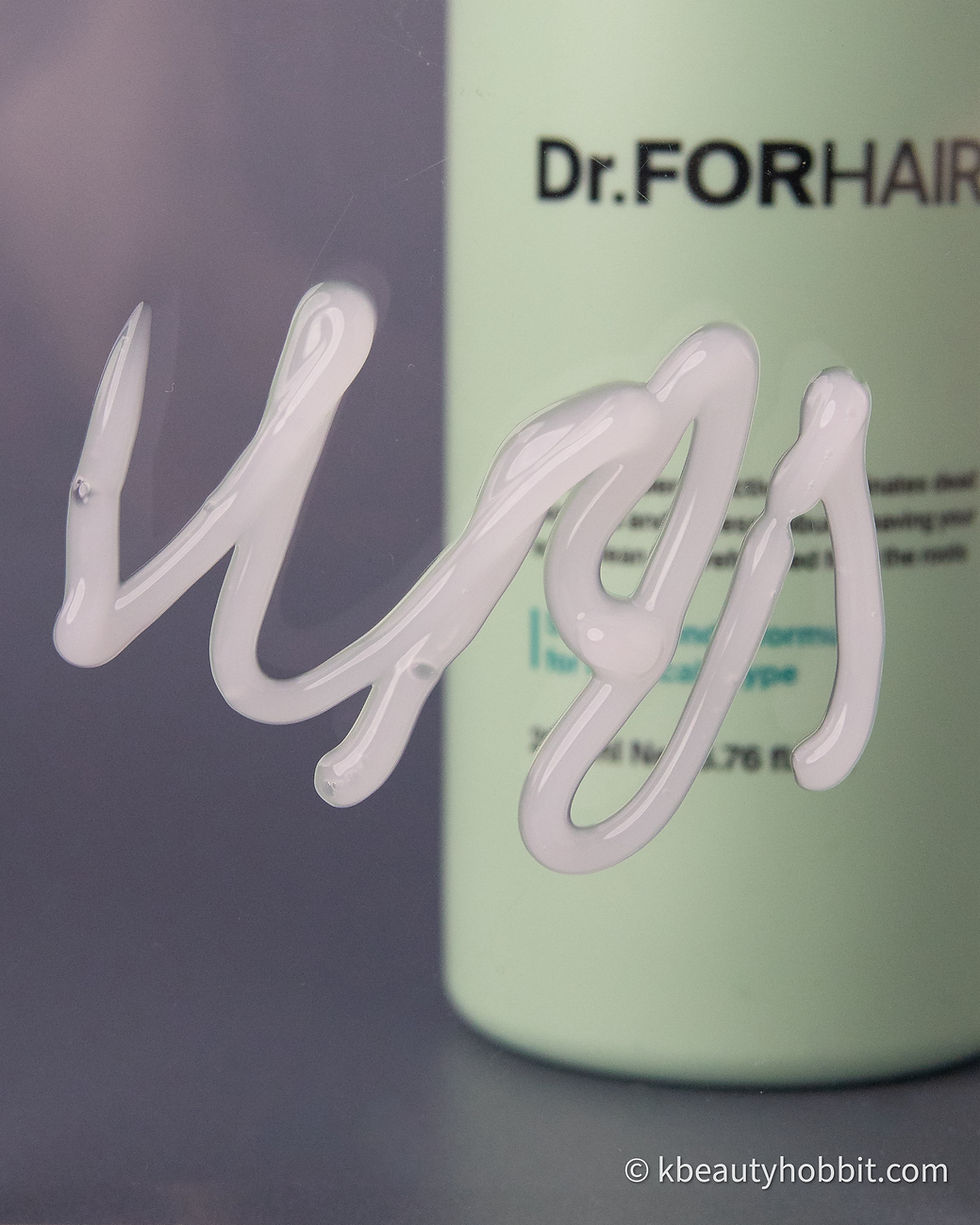 White cream on glass in front of a light green Dr.FORHAIR bottle. Blurred text in background, calm mood.
