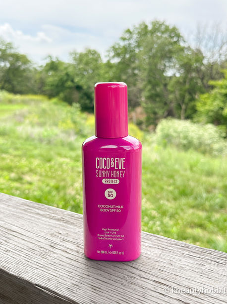 Bright pink Coco & Eve Sunny Honey Protect SPF 50 body sunscreen bottle on a wooden table with blurred green trees behind.