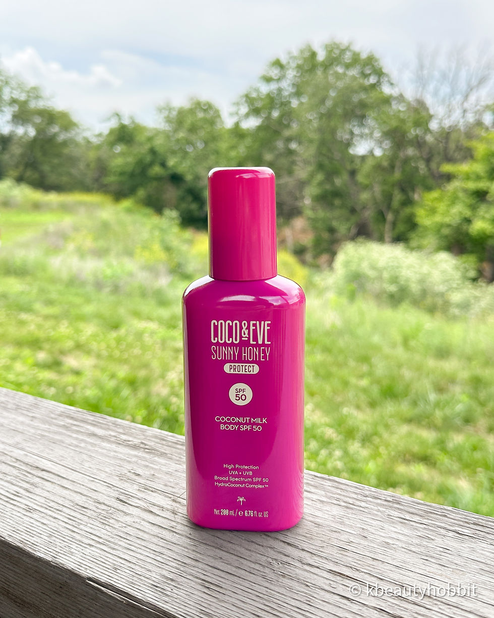 Bright pink Coco & Eve Sunny Honey Protect SPF 50 body sunscreen bottle on a wooden table with blurred green trees behind.