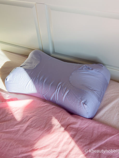 Sunlit bed with a lavender Omnia pillow on pink bedding, against a white headboard; watermark kbeautyhobbit in corner.