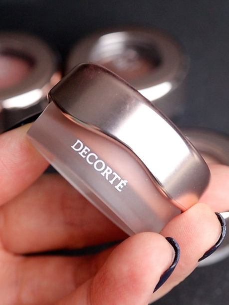 Hand holding a silver DECORTE eyeshadow pot, with blurred matching pieces on a dark background and kbeautyhobbit watermark.