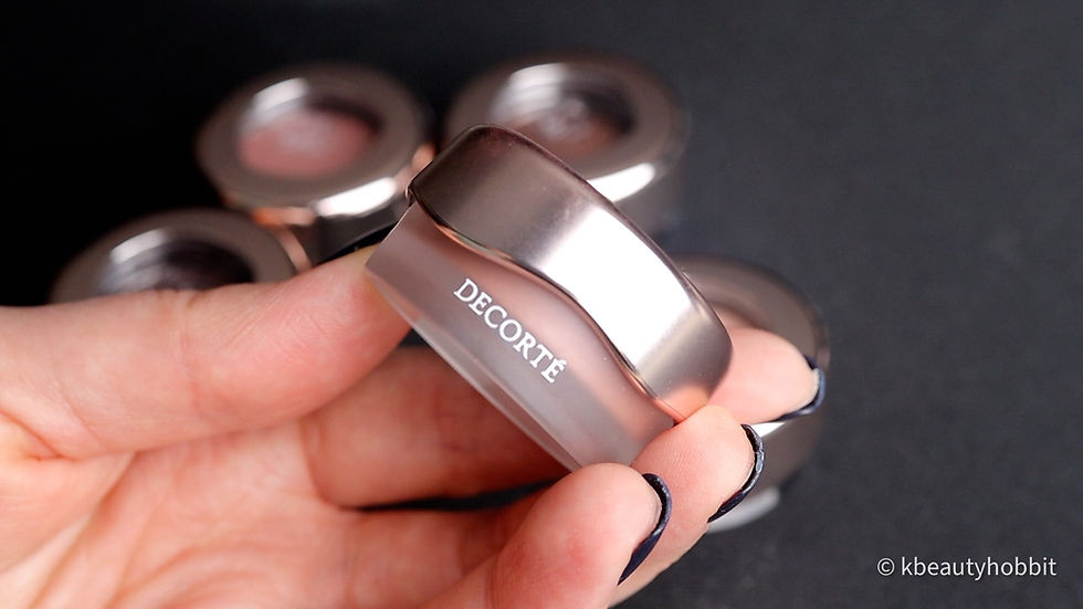 Hand holding a silver DECORTE eyeshadow pot, with blurred matching pieces on a dark background and kbeautyhobbit watermark.