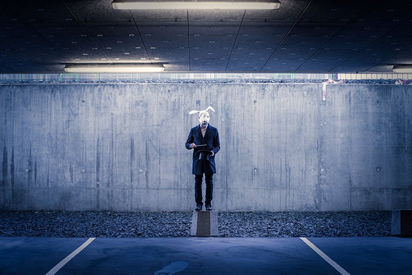 A man wearing a mask of a rabbit in front of a concrete wall, simulating a magic trick with a hat.