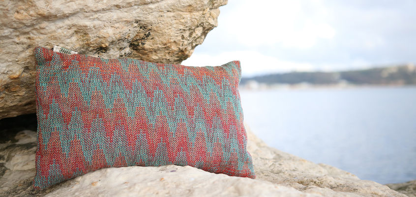 A handwoven lambswool merino cushion sits on top of a rock and beside another with the sea blurred in the background. The cushion is woven in the echo weave technique and features reds, greens and turquoises colours inspired by the Western Australian coastline. Handwoven by Sarah Affleck in Edinburgh Scotland using a 32 shaft Megado dobby loom.