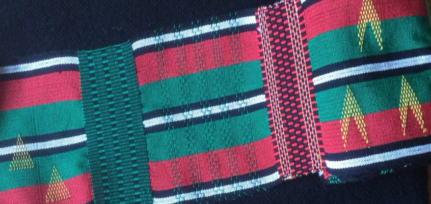 Detail from a roll of narrow warp and weft faced Kente style in red, green black and white mercerised cotton with yellow inlay geometric patterns shown on a black plain weave wool background