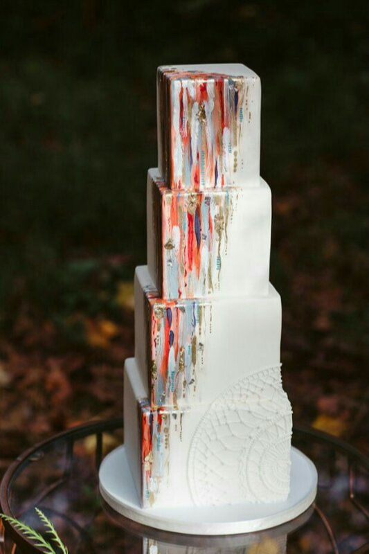 Este é um bolo de casamento de quatro andares com formato quadrado, base branca lisa e decoração artística de pinceladas coloridas (brushstrokes) com detalhes que parecem folha de ouro e um delicado relevo em renda na base.