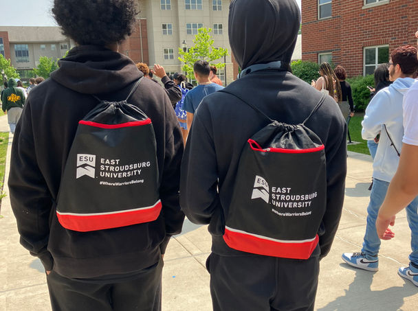 Two students wearing black and red East Stroudsburg University cinch bags look at brick buildings.