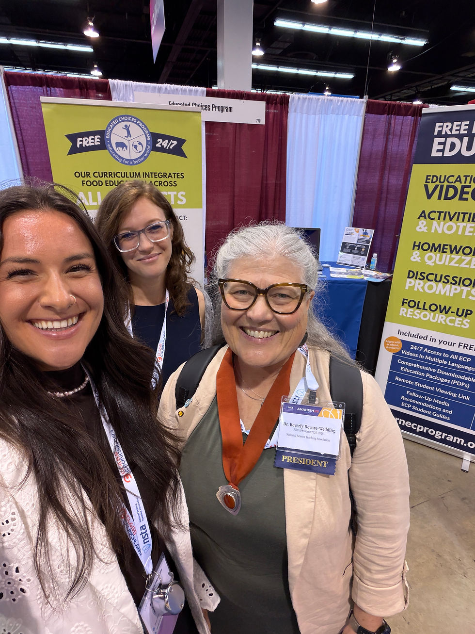 Three women smiling at a conference booth with banners about education. One wears a "PRESIDENT" badge. The setting is lively and colorful.