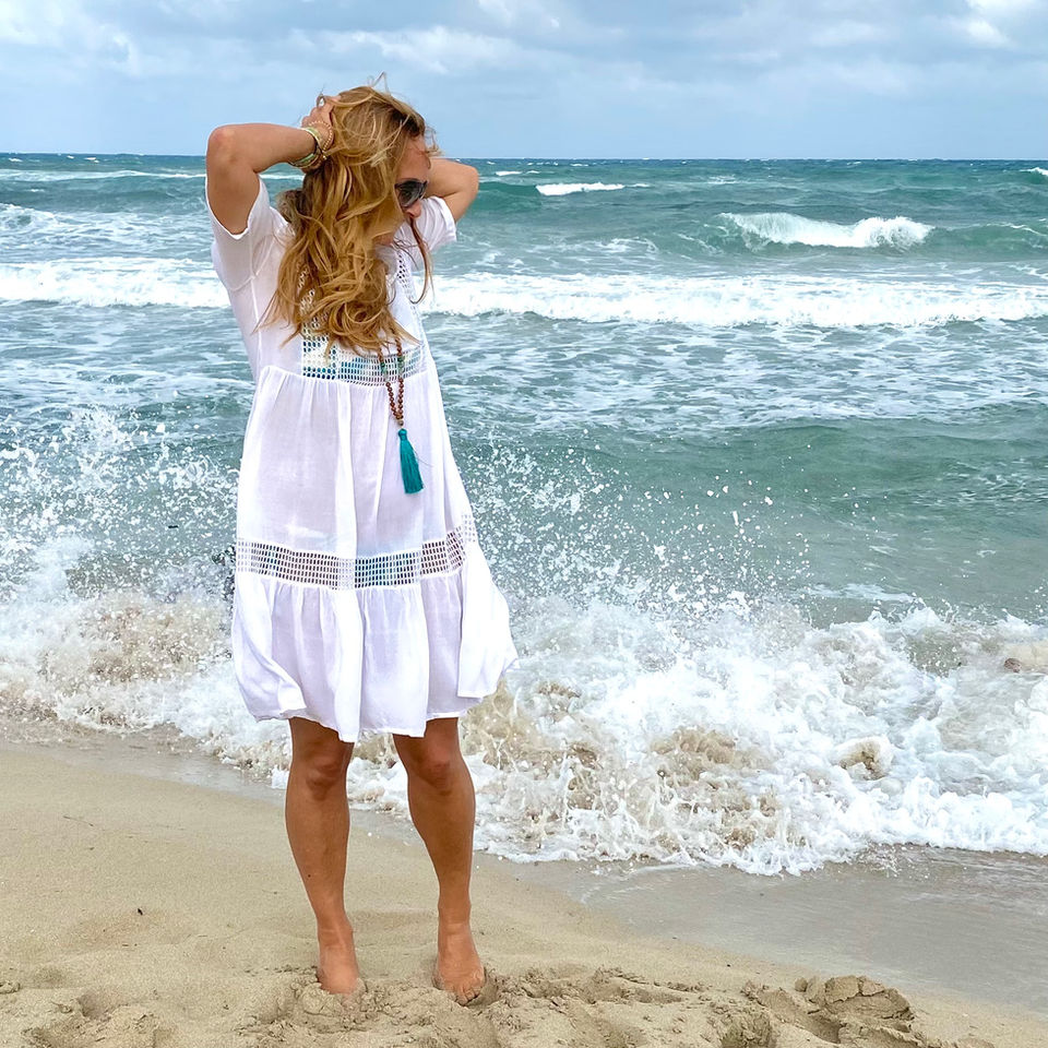 blond woman standing on a beach in Ibiza with the waves crashing wearing a white dress and a mala necklace