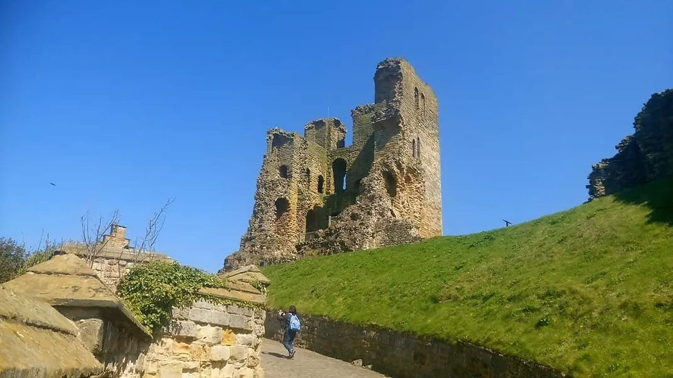 A person walks towards an ancient stone tower ruin under a clear blue sky, surrounded by green grass and stone walls.