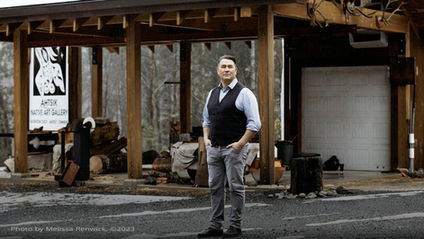 A man standing confidently in front of a wooden structure with a sign reading "AHTSIK Native Art Gallery" and "Gordon Dick, Artist/Owner." The setting includes logs, tools, and a garage door, with a forested background. The man is dressed in a blue shirt, black vest, and gray pants. Photo by Melissa Renwick, ©2023.