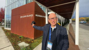 Phil Grabsky standing outside the San Juan Islands Museum of Art (SJIMA) in Friday Harbor, pointing toward the museum’s rust-colored exterior sign during his October 2025 visit.