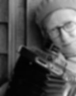 Black-and-white self-portrait: Imogen Cunningham leans into the frame beside a weathered wooden wall, wearing round glasses and a knit cap, with the bellows and lens of a large-format camera filling the lower half of the picture.