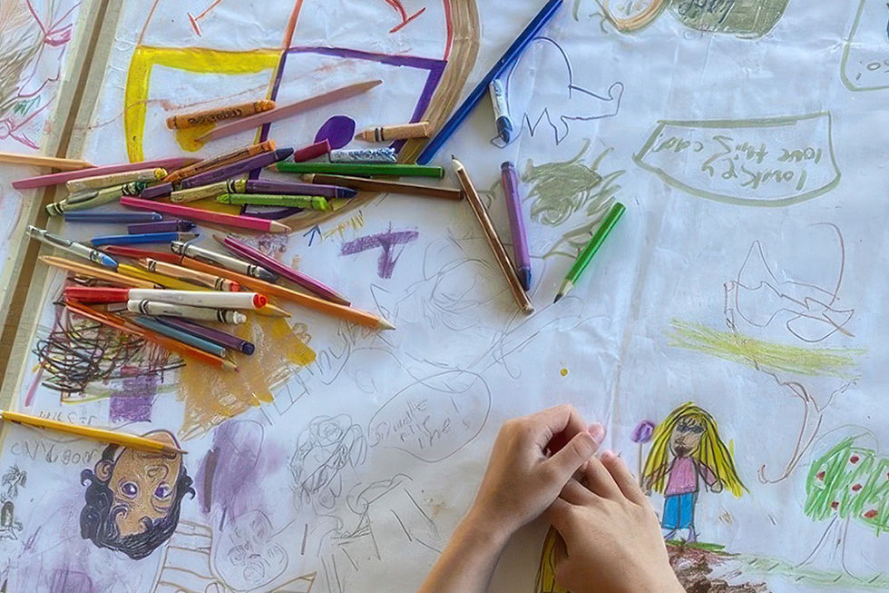 Child's hands drawing colorful comic-style characters and scenes on paper with crayons and colored pencils — a multimedia zine Family Art Days class at SJIMA on San Juan Island.