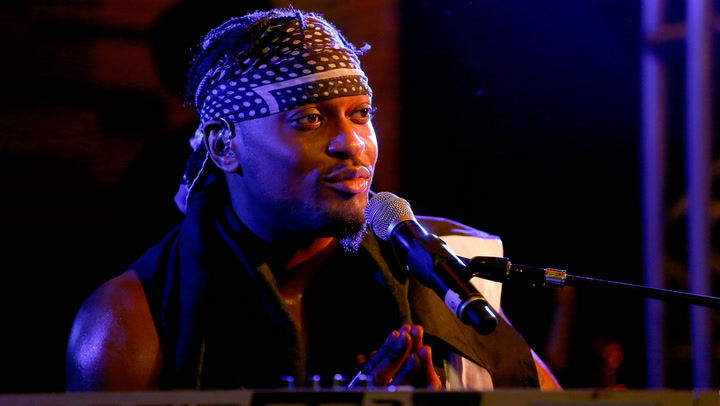 D'Angelo sits at a keyboard, smirk on his face, headband around his head, bare arms on display