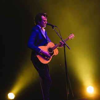 Paul Dempsey standing alone in a white spotlight, singing into a microphone while strumming an acoustic guitar before the glorious QPAC Concert Hall