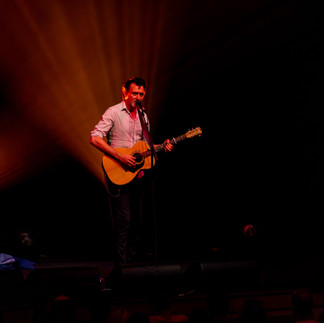 Paul Dempsey standing alone in a white spotlight, singing into a microphone while strumming an acoustic guitar before the glorious QPAC Concert Hall