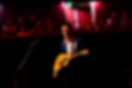 Paul Dempsey standing alone in a white spotlight, singing into a microphone while strumming an acoustic guitar.