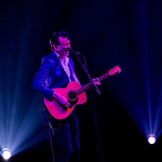 Paul Dempsey standing alone in a white spotlight, singing into a microphone while strumming an acoustic guitar before the glorious QPAC Concert Hall