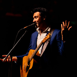 Paul Dempsey standing alone in a white spotlight, singing into a microphone while strumming an acoustic guitar before the glorious QPAC Concert Hall