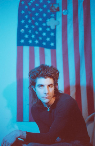 J Mahon in front of a large American flag, gazing into the camera with a calm, introspective expression.
