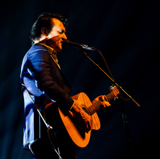 Paul Dempsey standing alone in a white spotlight, singing into a microphone while strumming an acoustic guitar before the glorious QPAC Concert Hall