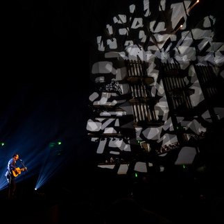 Paul Dempsey standing alone in a white spotlight, singing into a microphone while strumming an acoustic guitar before the glorious QPAC Concert Hall