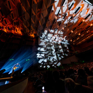 Paul Dempsey standing alone in a white spotlight, singing into a microphone while strumming an acoustic guitar before the glorious QPAC Concert Hall