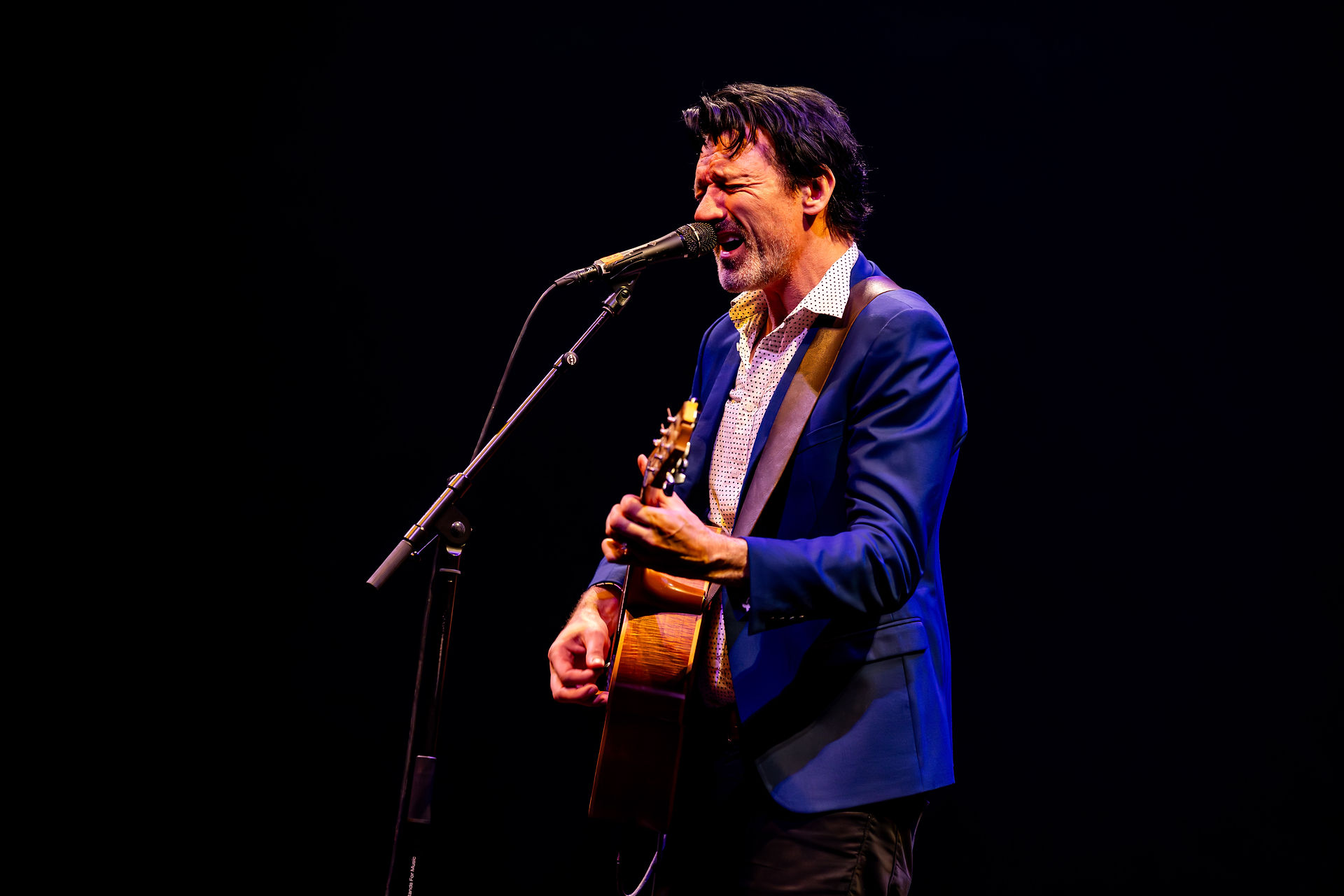 Paul Dempsey standing alone in a white spotlight, singing into a microphone while strumming an acoustic guitar before the glorious QPAC Concert Hall
