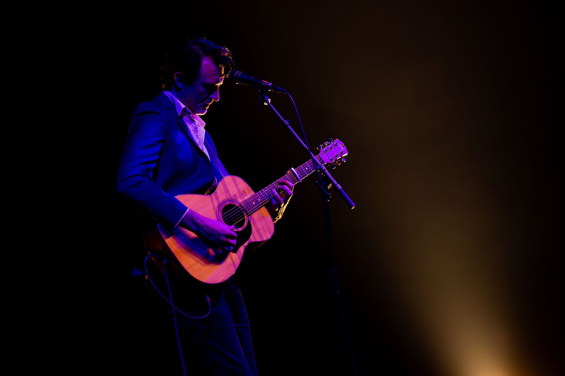 Paul Dempsey standing alone in a white spotlight, singing into a microphone while strumming an acoustic guitar before the glorious QPAC Concert Hall
