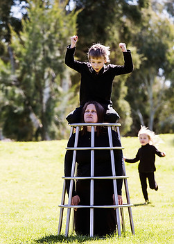image: the artwork 'structure' (2018) by contemporary jeweller Tiffany Parbs, made of a series of images of two children playing in the park on a stainless steel climbing frame being worn by their kneeling mother. This image shows the young girl running towards the mother/structure with the boy kneeling on top of the frame with the mother's head between his knees and his fists rasied © Tiffany Parbs, all rights reserved.