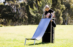 image: slope (2018), an artwork showing a series of images of a boy and a girl playing in the park on a three quarter sized wearable stainless steel slippery dip worn by their mother. It is a wearable neckpiece, with the mothers head poking through a hole in the top surface of the slide. The children are negotiating around the mothers head at the top of the ladder as they prepare to slide down © Tiffany Parbs, all rights reserved.