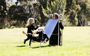 image: slope (2018), an artwork showing a series of images of a boy and a girl playing in the park on a three quarter sized wearable stainless steel slippery dip worn by their mother. It is a wearable neckpiece, with the mothers head poking through a hole in the top surface of the slide. The children are negotiating around the mothers head at the top of the ladder as they prepare to slide down © Tiffany Parbs, all rights reserved.
