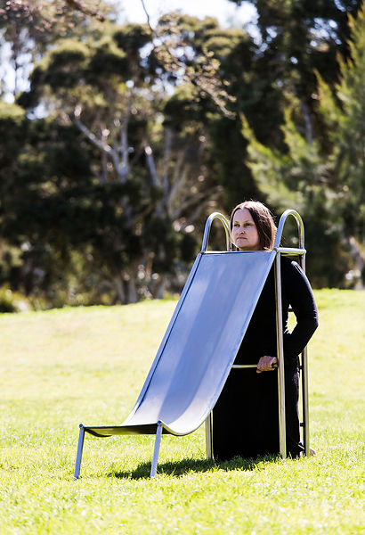 image: slope (2018), an artwork showing a series of images of a boy and a girl playing in the park on a three quarter sized wearable stainless steel slippery dip worn by their mother. In this image the woman is wearing the slide neckpiece, with her head poking through a hole in the top surface of the slide, the slide extended and balanced on the ground. The woman looks strong, alert and watchful, the children are not present in this particular image from the series. © Tiffany Parbs, all rights reserved.